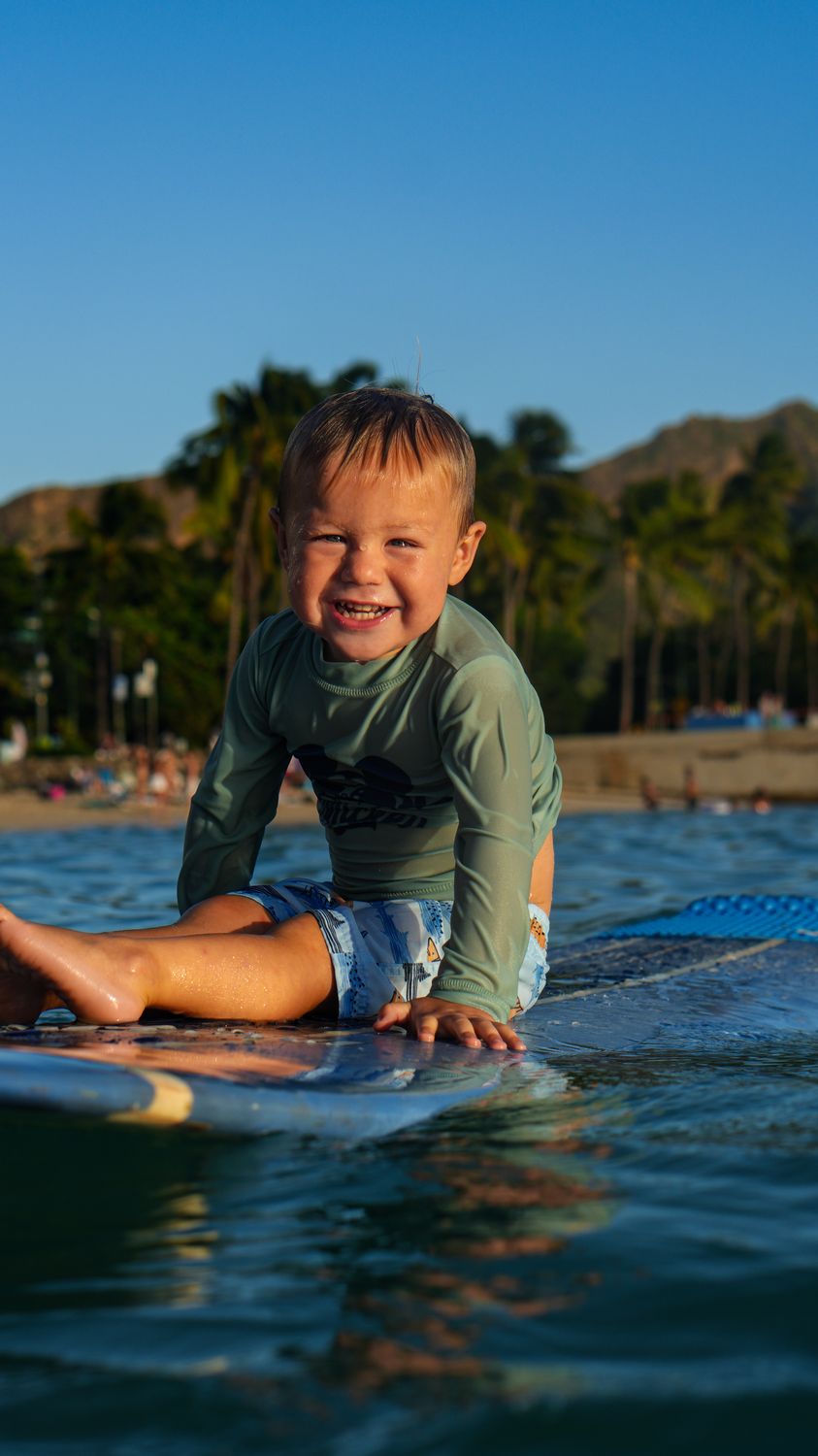 Toddler sitting on a surfboard at Waikiki, grinning at the camera