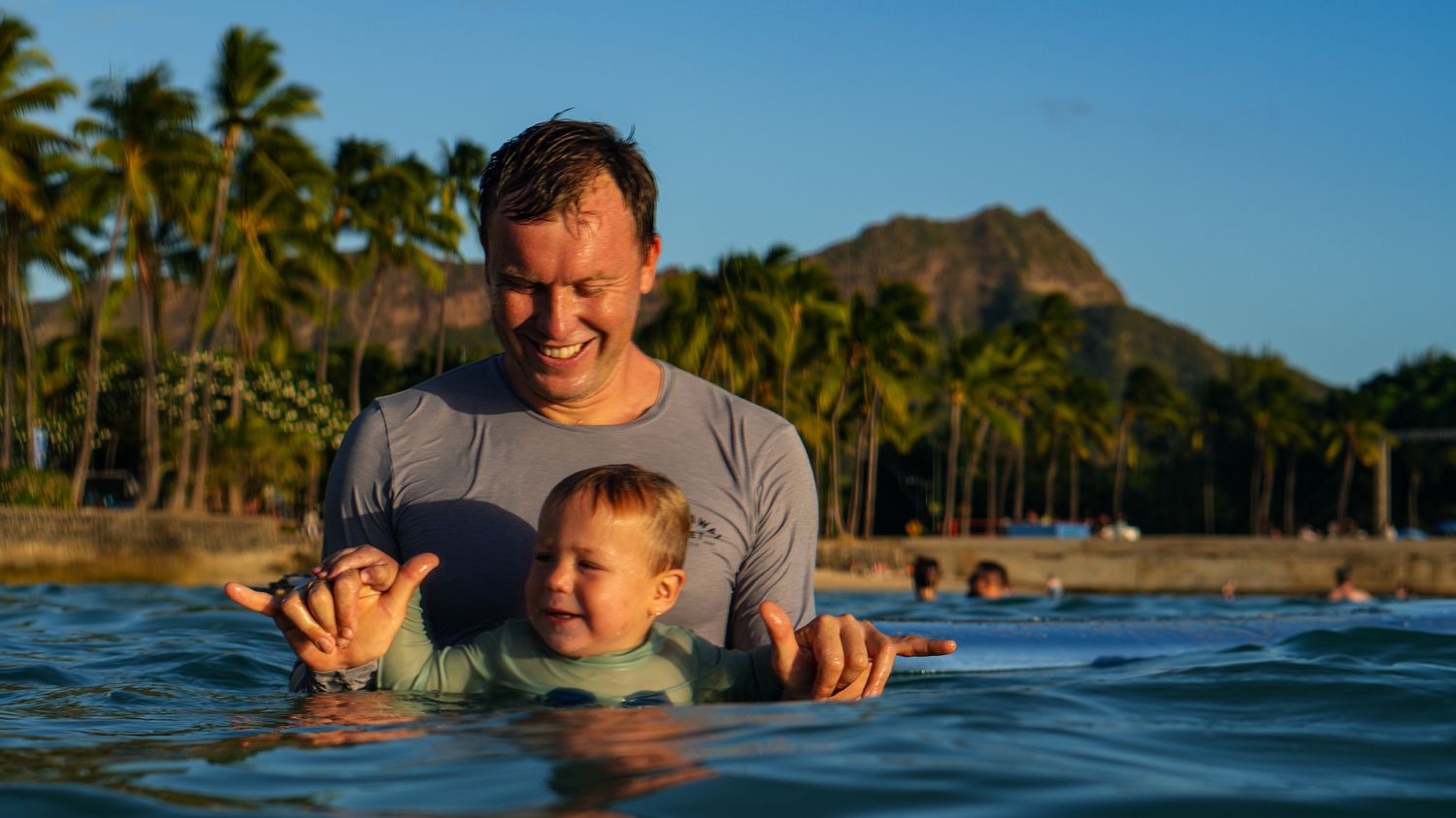 Dad and toddler throwing shakas in the ocean with Diamond Head behind
