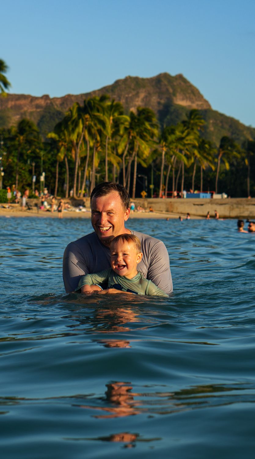 Dad holding his laughing toddler in the ocean at Waikiki with Diamond Head behind