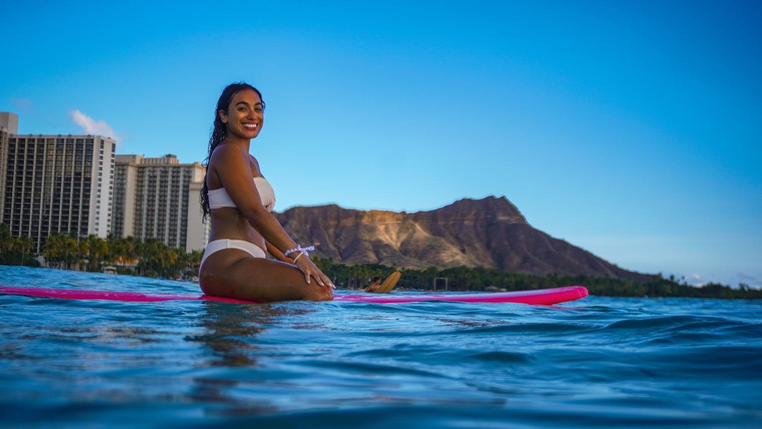 Woman in white bikini sitting on pink surfboard with Diamond Head behind