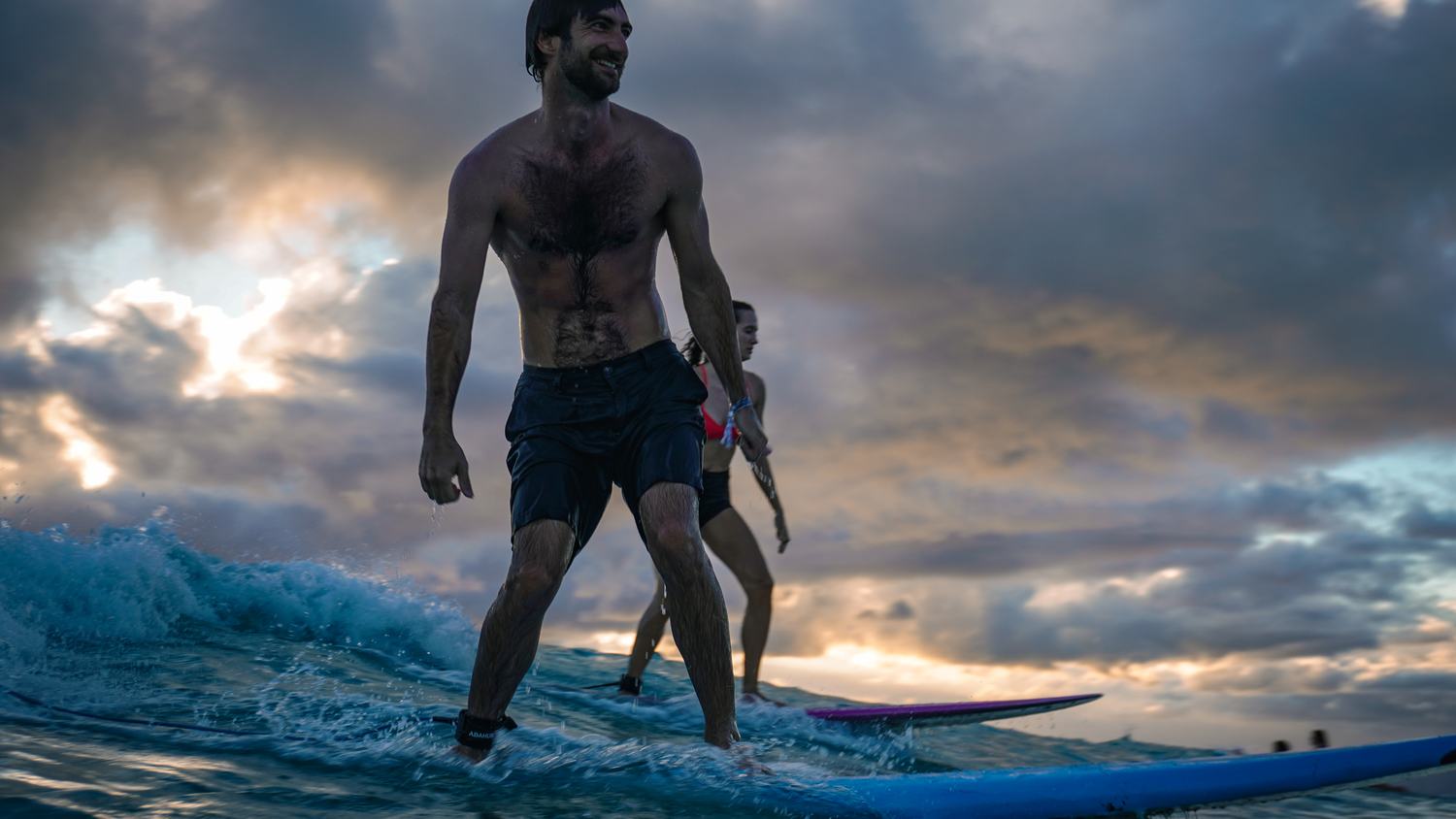 Man walking on a surfboard with a woman behind under dramatic clouds